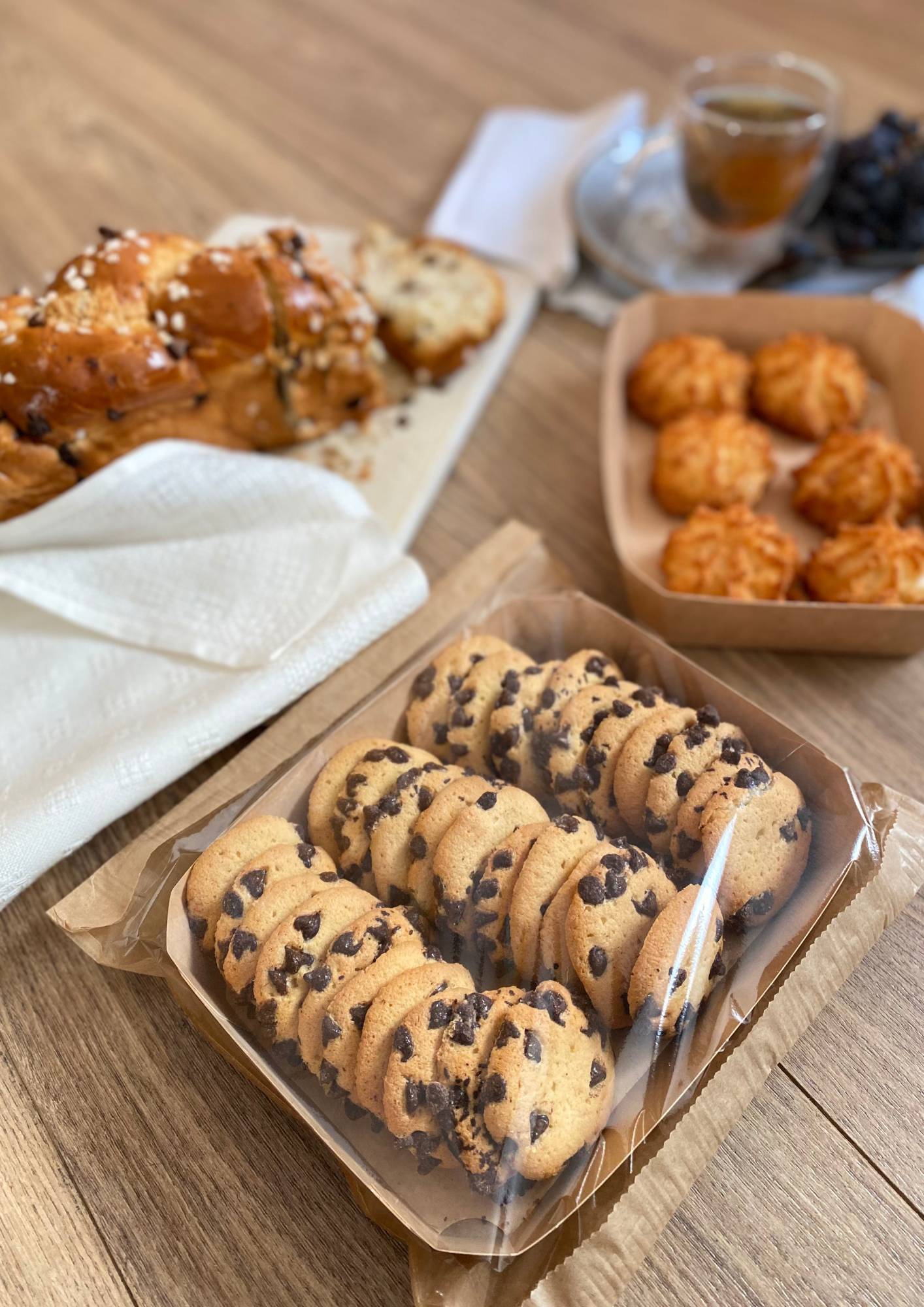 In the foreground a tray of chocolate chip cookies, in the background coconut macaroons, a braided loaf of bread, a cup of tea and grapes on a wooden table.