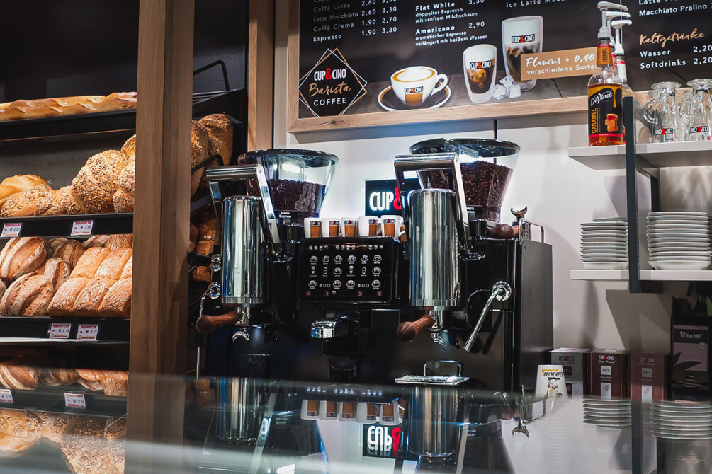 Eine Kaffeemaschine mit gestapelten Tassen steht auf der Theke einer Bäckerei, umgeben von Brotlaiben auf Regalen und einer Menütafel darüber. Im Vordergrund ist eine Glasvitrine zu sehen.