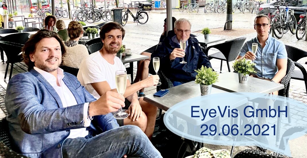 Vier Männer sitzen an einem Cafétisch im Freien, lächeln und stoßen mit einem Glas Champagner an. Die Szene ist entspannt und leger. Ein blauer Text überlagert EyeVis GmbH 29.06.2021. Im Hintergrund sind Fahrräder und Menschen zu sehen.